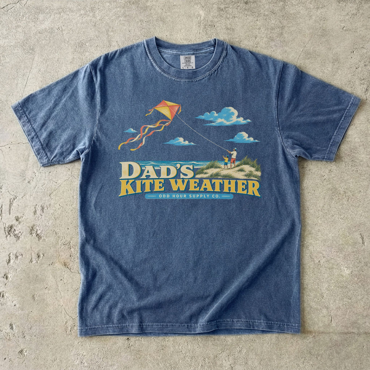 Comfort Colors 1717 T-Shirt in Denim mit Papa und Kind in Stranddünen, orange-gelbem Flugdrachen, Meer, Wolken und Dad’s Kite Weather-Schrift.