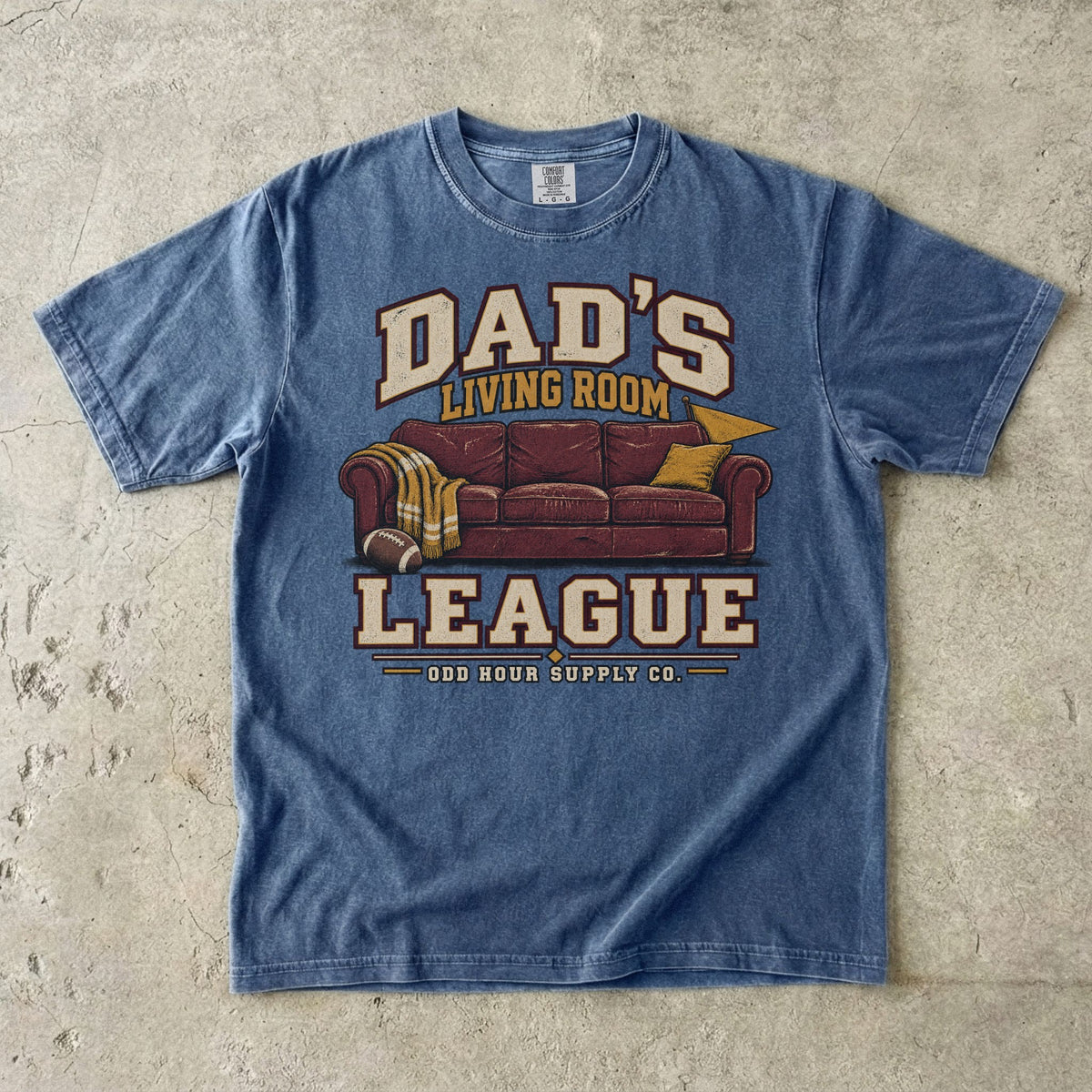 Comfort Colors 1717 T-Shirt in Denim mit rotbraunem Sofa, Football, gelber Decke und Dad’s Living Room League-Schrift.