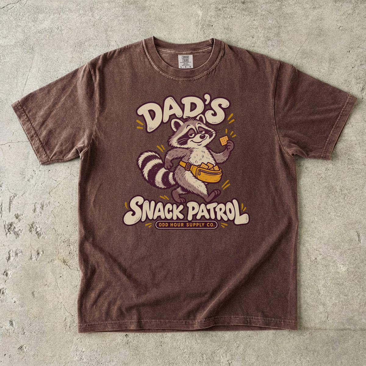 Comfort Colors 1717 T-Shirt in Espresso mit Waschbär, gelber Bauchtasche voller Cracker und cremefarbener Dad’s Snack Patrol-Schrift.