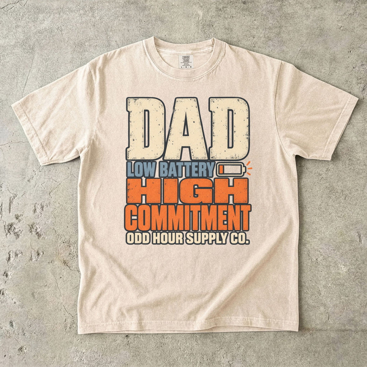 Comfort Colors 1717 T-Shirt in Ivory mit großer Dad Low Battery High Commitment-Schrift in Creme, Blau und Orange sowie fast leerem Batteriesymbol.