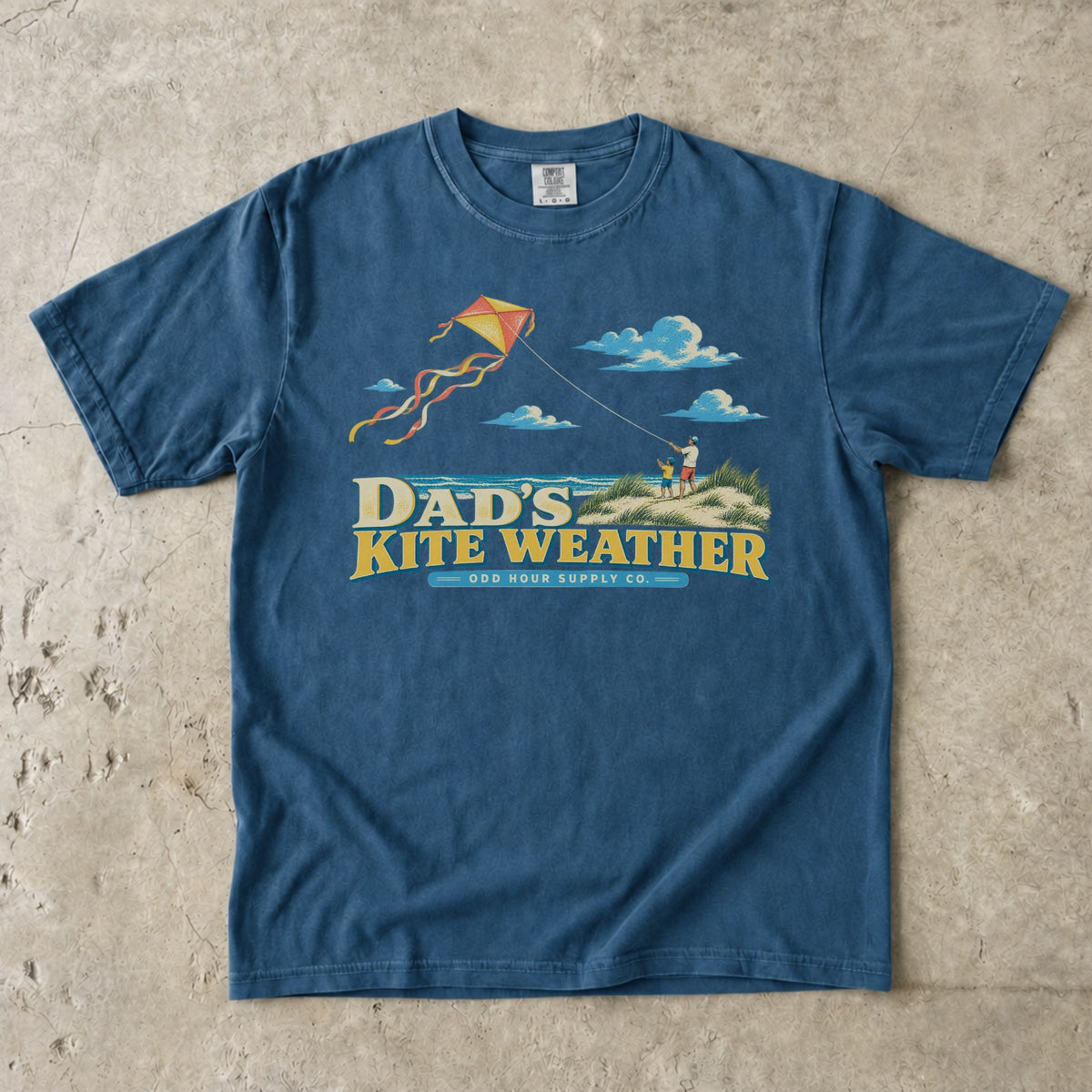 Comfort Colors 1717 T-Shirt in Midnight mit Papa und Kind in Stranddünen, orange-gelbem Flugdrachen, Meer, Wolken und Dad’s Kite Weather-Schrift.