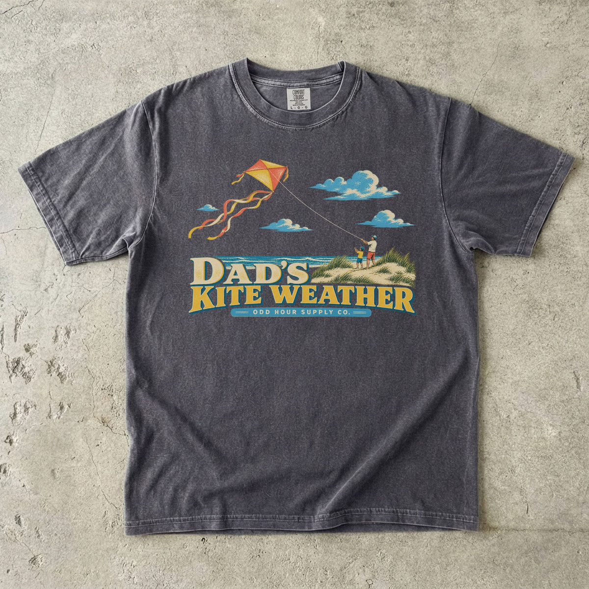Comfort Colors 1717 T-Shirt in Pepper mit Papa und Kind in Stranddünen, orange-gelbem Flugdrachen, Meer, Wolken und Dad’s Kite Weather-Schrift.