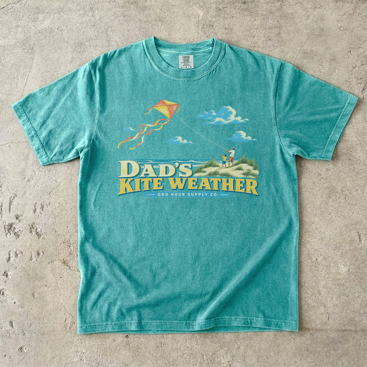 Comfort Colors 1717 T-Shirt in Seafoam mit Papa und Kind in Stranddünen, orange-gelbem Flugdrachen, Meer, Wolken und Dad’s Kite Weather-Schrift.