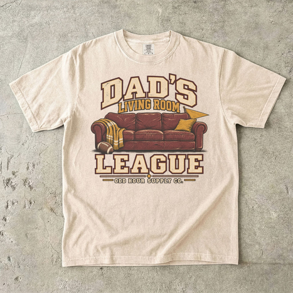 Comfort Colors 1717 T-Shirt in Ivory mit rotbraunem Sofa, Football, gelber Decke und Dad’s Living Room League-Schrift.