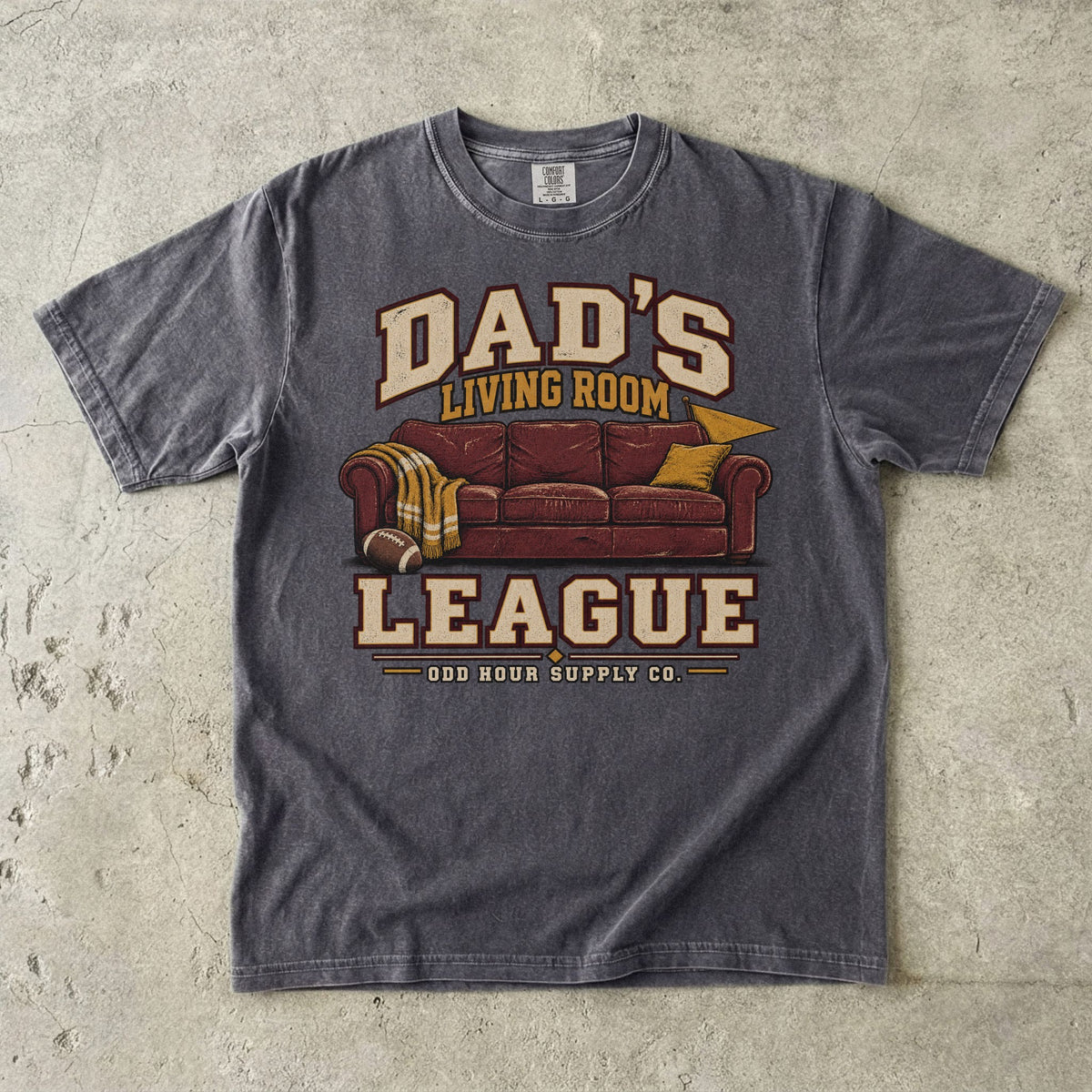 Comfort Colors 1717 T-Shirt in Pepper mit rotbraunem Sofa, Football, gelber Decke und Dad’s Living Room League-Schrift.