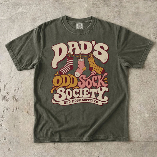 Comfort Colors 1717 T-Shirt in Sage mit drei gemusterten Socken an einer Wäscheleine und Dad’s Odd Sock Society-Schrift in Creme, Rosa und Senfgelb.