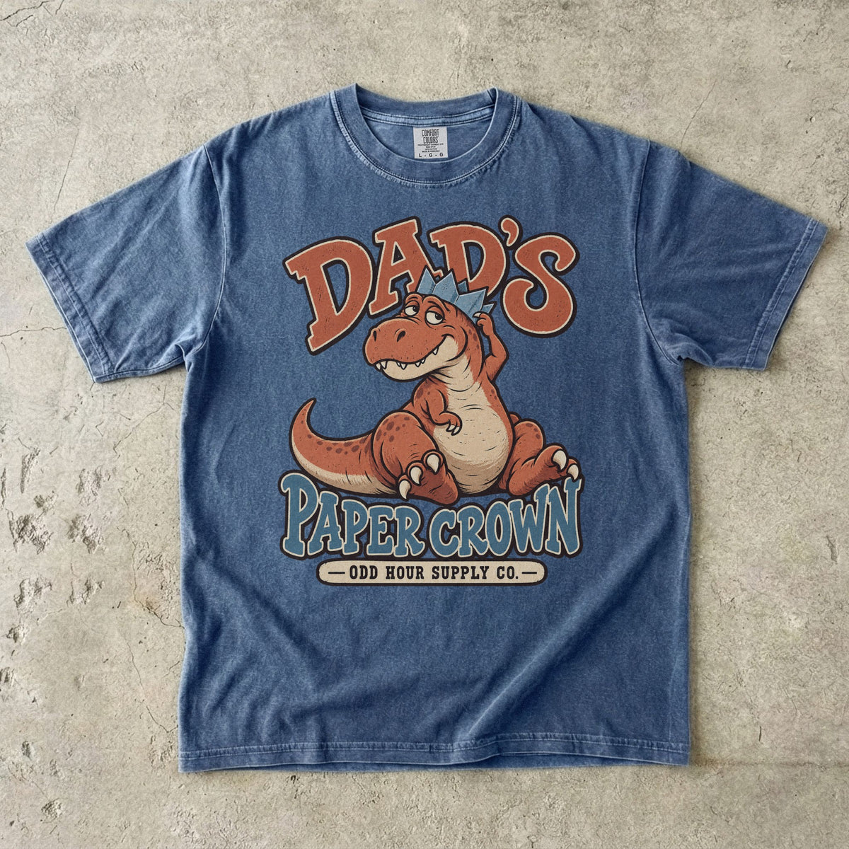 Comfort Colors 1717 T-Shirt in Denim mit sitzendem orangefarbenem Dinosaurier, blauer Papierkrone und Dad’s Paper Crown-Schrift.