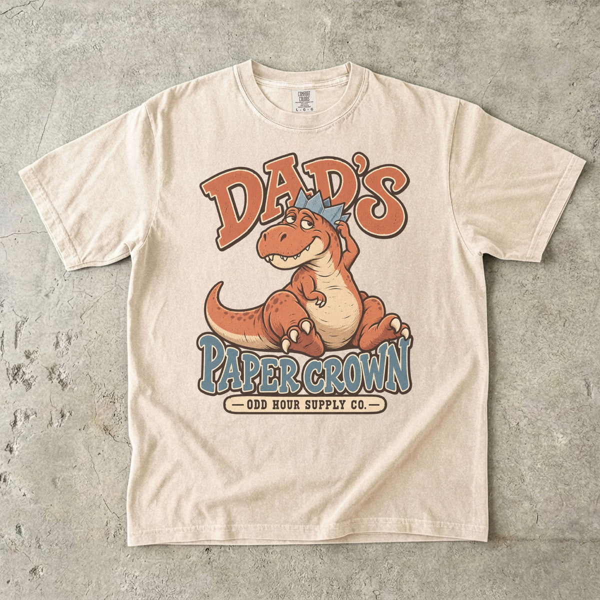 Comfort Colors 1717 T-Shirt in Ivory mit sitzendem orangefarbenem Dinosaurier, blauer Papierkrone und Dad’s Paper Crown-Schrift.