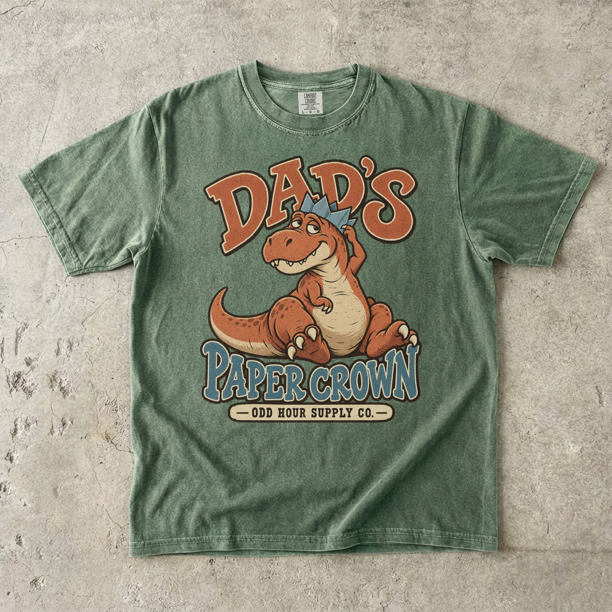 Comfort Colors 1717 T-Shirt in Moss mit sitzendem orangefarbenem Dinosaurier, blauer Papierkrone und Dad’s Paper Crown-Schrift.