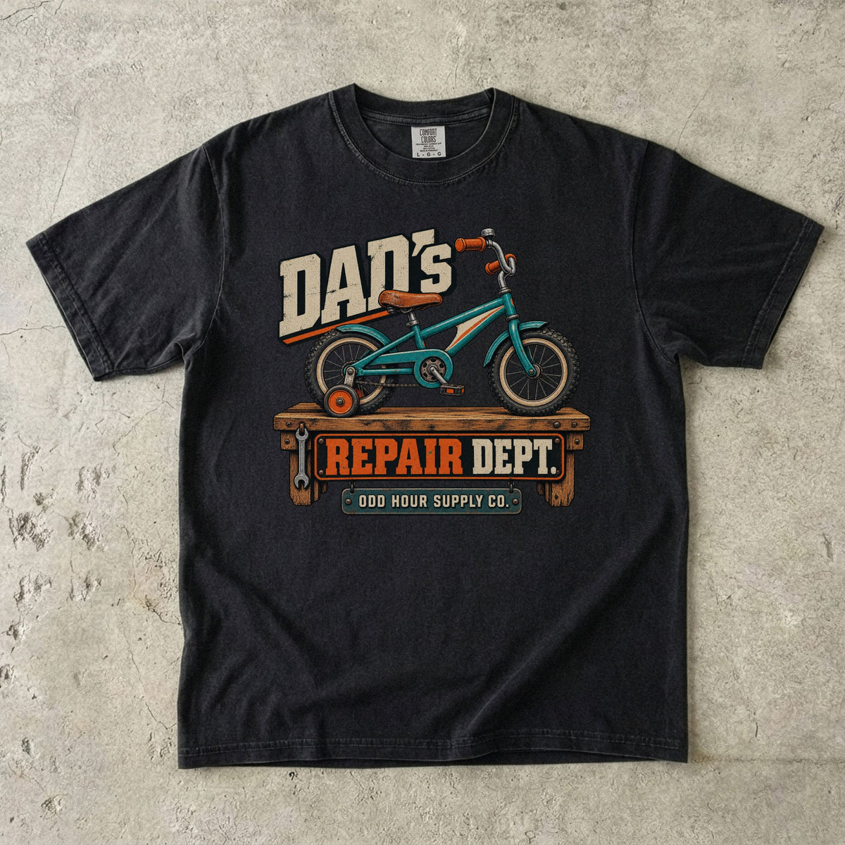 Comfort Colors 1717 T-Shirt in Black mit türkisfarbenem Kinderfahrrad mit Stützrad auf einer Werkbank und Dad’s Repair Dept.-Schrift.
