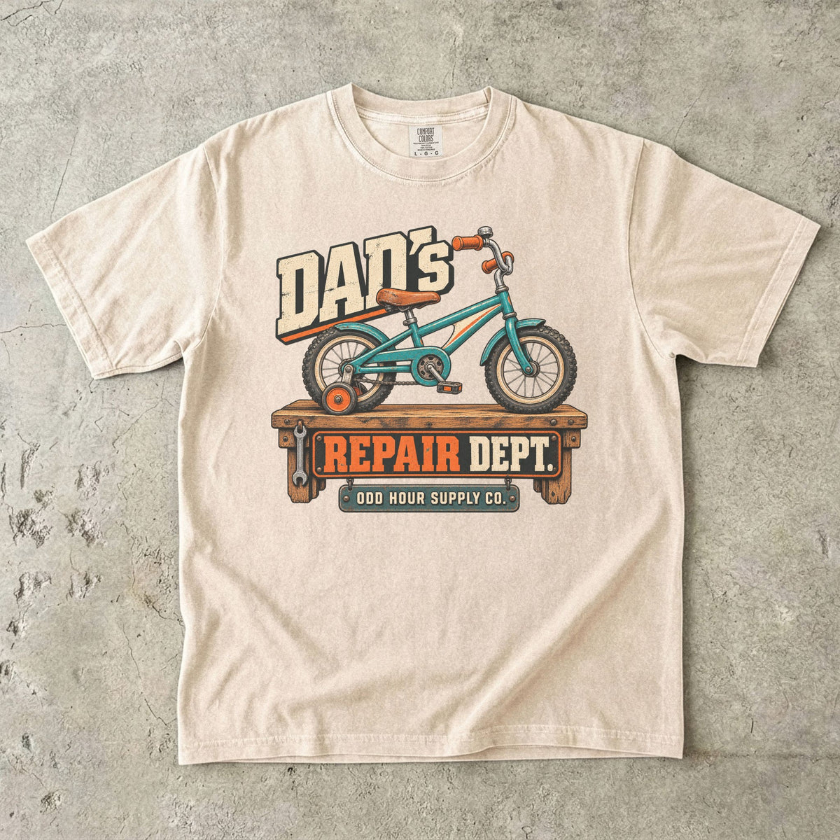 Comfort Colors 1717 T-Shirt in Ivory mit türkisfarbenem Kinderfahrrad mit Stützrad auf einer Werkbank und Dad’s Repair Dept.-Schrift.