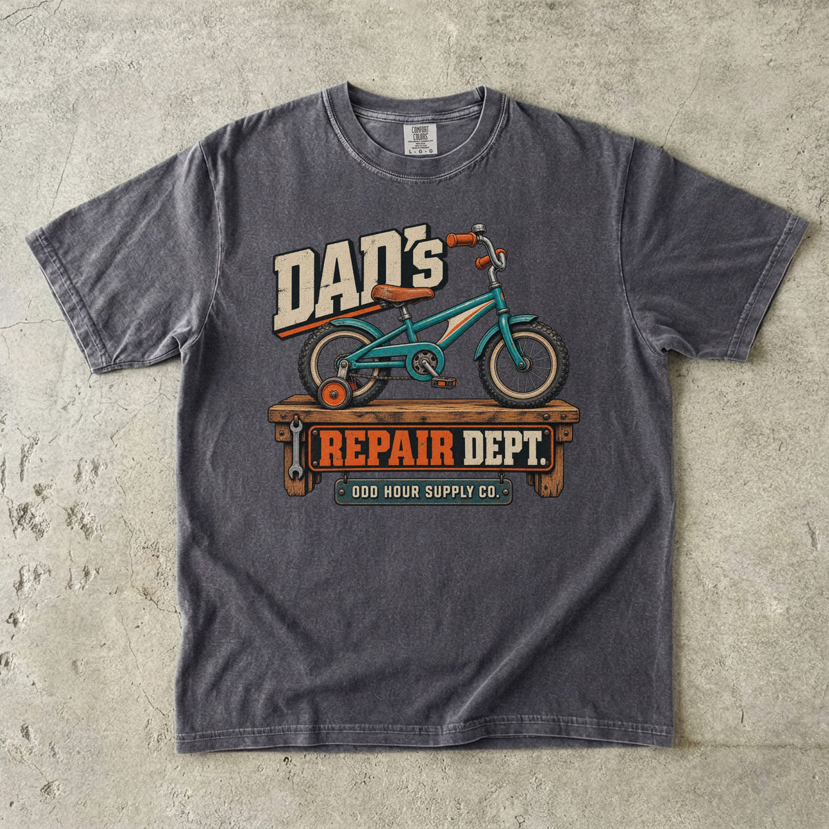 Comfort Colors 1717 T-Shirt in Pepper mit türkisfarbenem Kinderfahrrad mit Stützrad auf einer Werkbank und Dad’s Repair Dept.-Schrift.