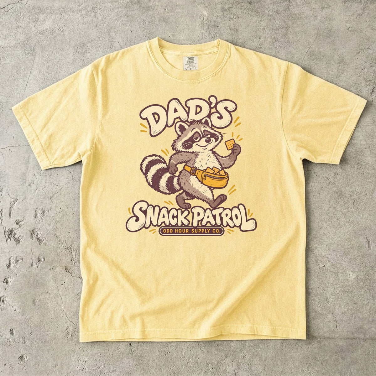 Comfort Colors 1717 T-Shirt in Butter mit Waschbär, gelber Bauchtasche voller Cracker und cremefarbener Dad’s Snack Patrol-Schrift.