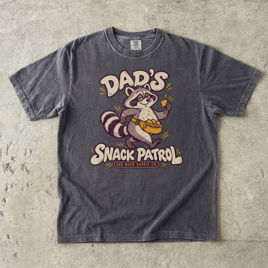 Comfort Colors 1717 T-Shirt in Pepper mit Waschbär, gelber Bauchtasche voller Cracker und cremefarbener Dad’s Snack Patrol-Schrift.