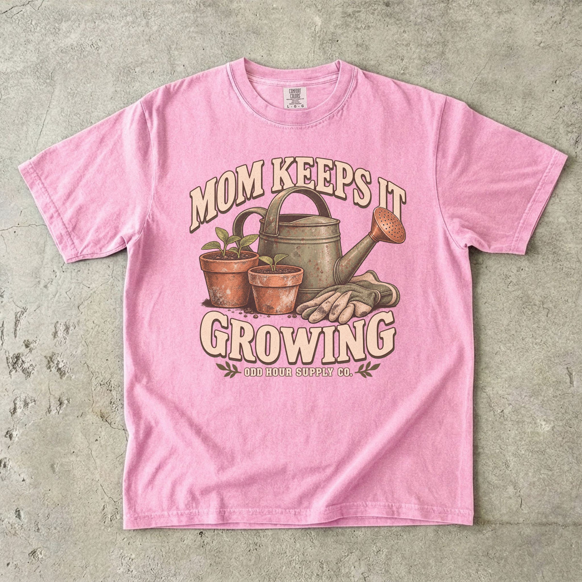 Comfort Colors 1717 T-Shirt in Blossom mit grüner Gießkanne, zwei Terrakottatöpfen mit Setzlingen, Gartenhandschuhen und Mom Keeps It Growing-Schrift.