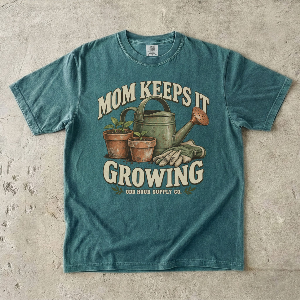 Comfort Colors 1717 T-Shirt in Blue Spruce mit grüner Gießkanne, zwei Terrakottatöpfen mit Setzlingen, Gartenhandschuhen und Mom Keeps It Growing-Schrift.