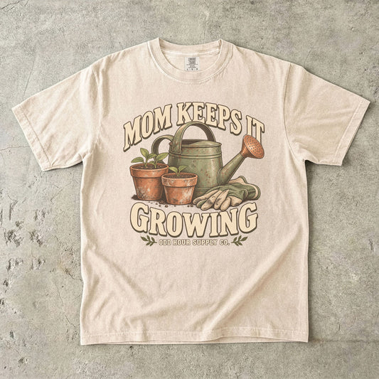 Comfort Colors 1717 T-Shirt in Ivory mit grüner Gießkanne, zwei Terrakottatöpfen mit Setzlingen, Gartenhandschuhen und Mom Keeps It Growing-Schrift.