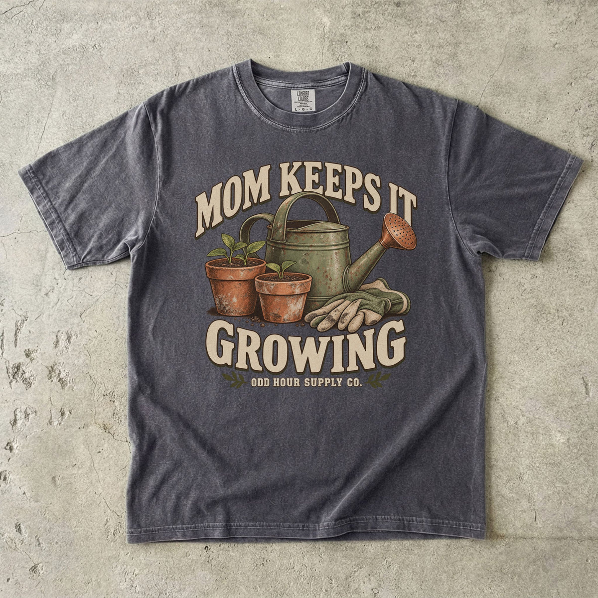 Comfort Colors 1717 T-Shirt in Pepper mit grüner Gießkanne, zwei Terrakottatöpfen mit Setzlingen, Gartenhandschuhen und Mom Keeps It Growing-Schrift.