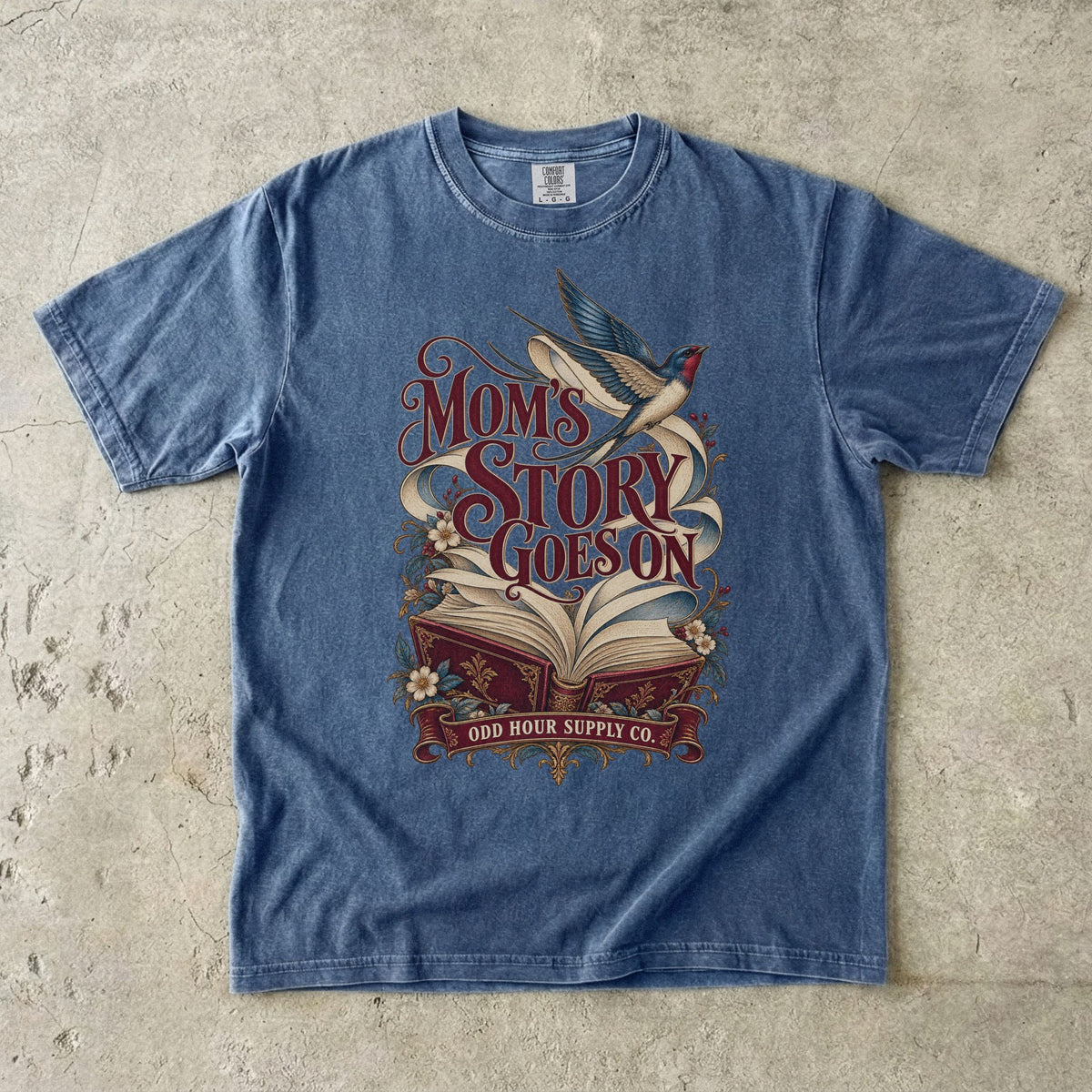 Comfort Colors 1717 T-Shirt in Denim mit offenem rotem Buch, geschwungenen Seiten, fliegender Schwalbe, Blumen und Mom’s Story Goes On-Schrift.