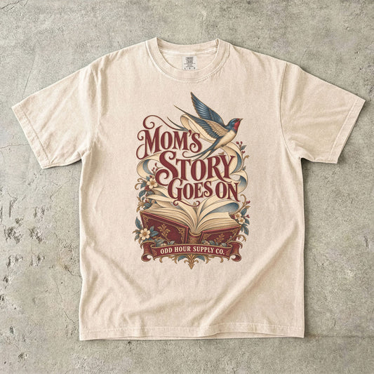 Comfort Colors 1717 T-Shirt in Ivory mit offenem rotem Buch, geschwungenen Seiten, fliegender Schwalbe, Blumen und Mom’s Story Goes On-Schrift.